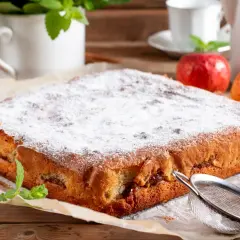 Puszyste ciasto bez tłuszczu, posypane cukrem pudrem, z jabłkami i cynamonem. Obok jabłka, mięta i sitko.