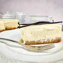 Kawałek puszystego new york cheesecake z wanilią na kruchym spodzie, udekorowany laskami wanilii. Obok widoczne inne serniki i filiżanka.