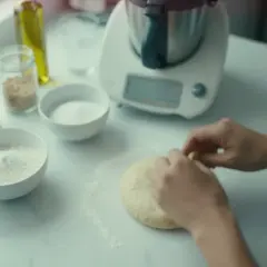 Ręce formują puszyste ciasto na pizzę na blacie. Obok miski z mąką, cukrem i nasionami.