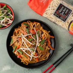 Pyszny makaron chiński z kurczakiem, papryką i kiełkami fasoli mung, gotowy do spożycia. Obok opakowanie makaronu Chow Mein.