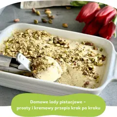 Kremowe lody pistacjowe w białej formie, posypane orzechami. Obok czerwone tulipany.