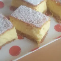 Kawałki puszystego sernika bez sera, posypane cukrem pudrem, na talerzu w czerwone kropki.