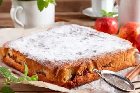 Puszyste ciasto bez tłuszczu, posypane cukrem pudrem, z jabłkami i cynamonem. Obok jabłka, mięta i sitko.