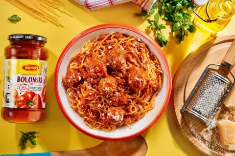 Pyszne spaghetti bolognese z klopsikami w sosie pomidorowym, obok słoik sosu i tarka z parmezanem.