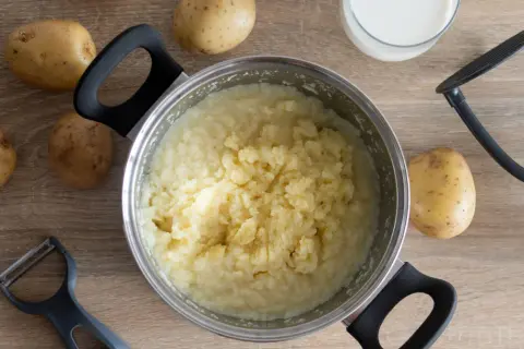 Gotujesz ziemniaki, by sprawdzić, czy są lekkostrawne. Obok garnka leżą surowe ziemniaki, mleko, tłuczek i obieraczka.