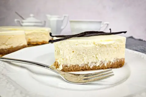 Kawałek puszystego new york cheesecake z wanilią na kruchym spodzie, udekorowany laskami wanilii. Obok widoczne inne serniki i filiżanka.