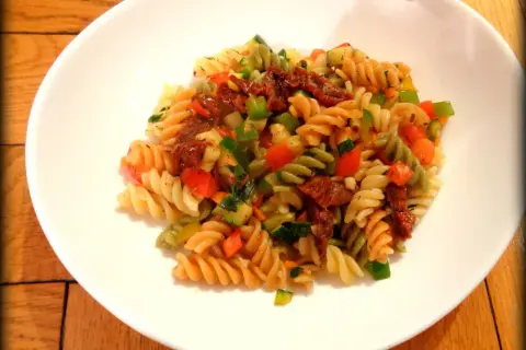 Kolorowy makaron fusilli z suszonymi pomidorami, cukinią, papryką i natką pietruszki. Pyszny makaron z warzywami na białym talerzu.