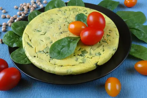 Puszysty omlet z ciecierzycy ze szpinakiem, ozdobiony pomidorkami koktajlowymi i świeżym szpinakiem.