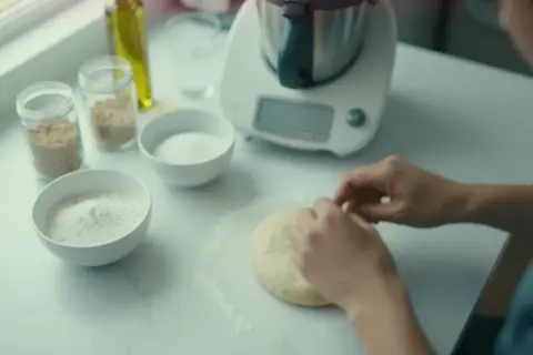 Ręce formują puszyste ciasto na pizzę na blacie. Obok miski z mąką, cukrem i nasionami.
