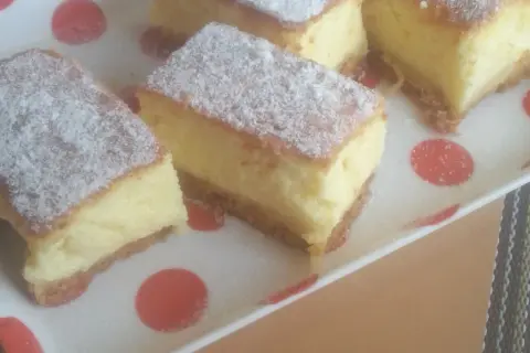 Kawałki puszystego sernika bez sera, posypane cukrem pudrem, na talerzu w czerwone kropki.
