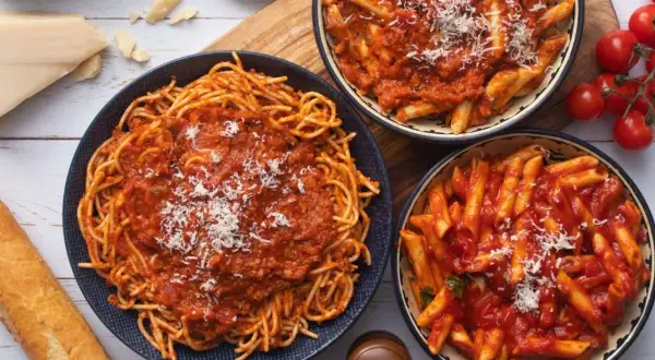 Pyszny obiad bez mięsa: spaghetti i penne w sosie pomidorowym, posypane parmezanem. Obok bagietka i pomidorki.