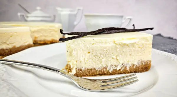 Kawałek puszystego new york cheesecake z wanilią na kruchym spodzie, udekorowany laskami wanilii. Obok widoczne inne serniki i filiżanka.