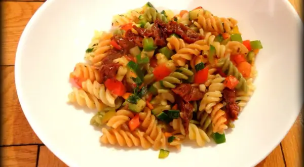 Kolorowy makaron fusilli z suszonymi pomidorami, cukinią, papryką i natką pietruszki. Pyszny makaron z warzywami na białym talerzu.