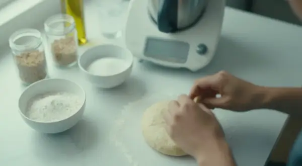 Ręce formują puszyste ciasto na pizzę na blacie. Obok miski z mąką, cukrem i nasionami.