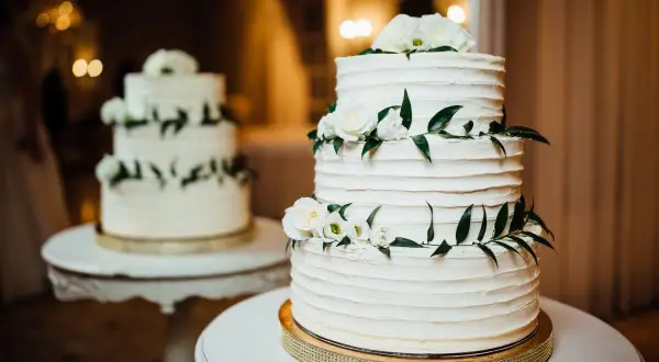 Elegancki tort piętrowy na stelażu, ozdobiony białymi różami i zielonymi liśćmi. Idealny pomysł, jak zrobić tort piętrowy na stelażu na specjalną okazję.
