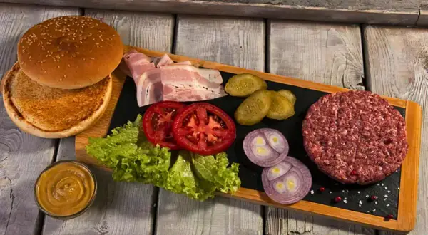 Wszystko, co do burgera: bułka, surowe mięso, boczek, pomidory, sałata, ogórki, cebula i musztarda.