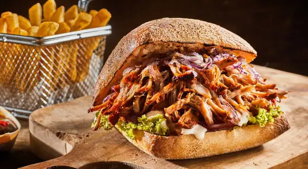 Soczysty pulled pork w bułce, z czerwoną cebulą i sałatą, obok frytki. Idealny pulled pork przepis na obiad.