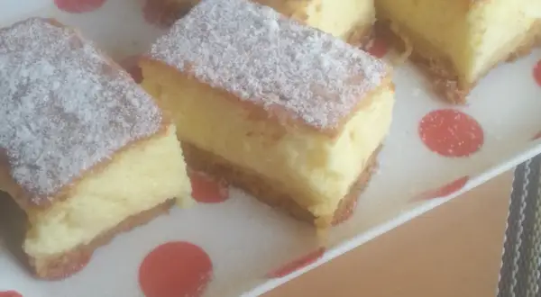 Kawałki puszystego sernika bez sera, posypane cukrem pudrem, na talerzu w czerwone kropki.