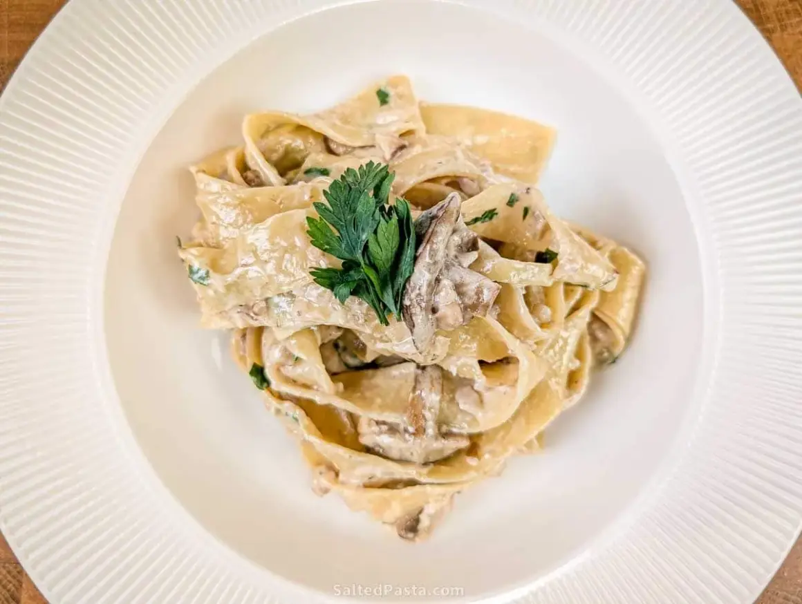 Pappardelle z kremowym sosem i kawałkami grzybów, udekorowane natką pietruszki. Pyszny makaron z grzybami.