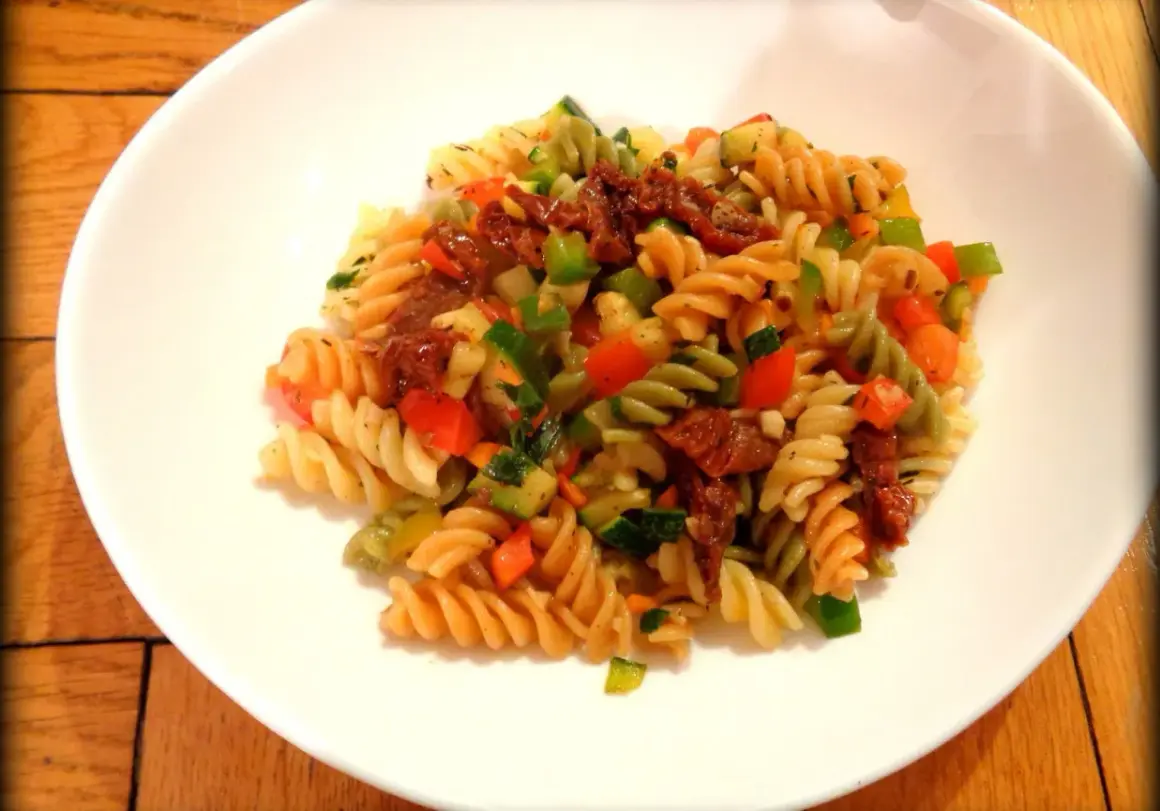 Kolorowy makaron fusilli z suszonymi pomidorami, cukinią, papryką i natką pietruszki. Pyszny makaron z warzywami na białym talerzu.