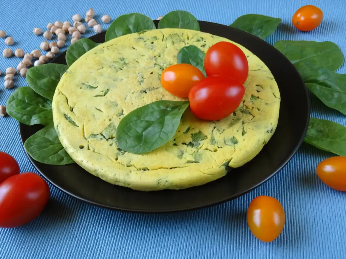 Puszysty omlet z ciecierzycy ze szpinakiem, ozdobiony pomidorkami koktajlowymi i świeżym szpinakiem.