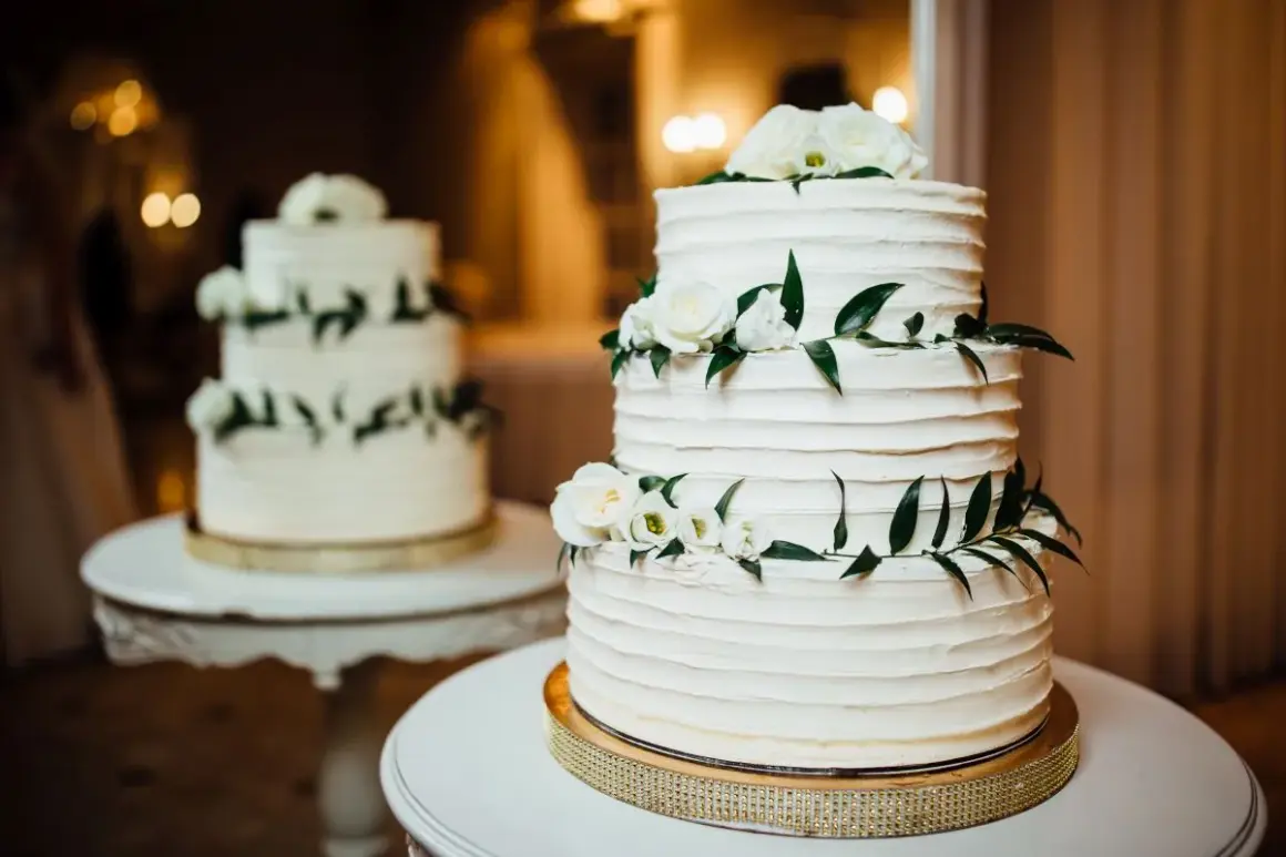 Elegancki tort piętrowy na stelażu, ozdobiony białymi różami i zielonymi liśćmi. Idealny pomysł, jak zrobić tort piętrowy na stelażu na specjalną okazję.