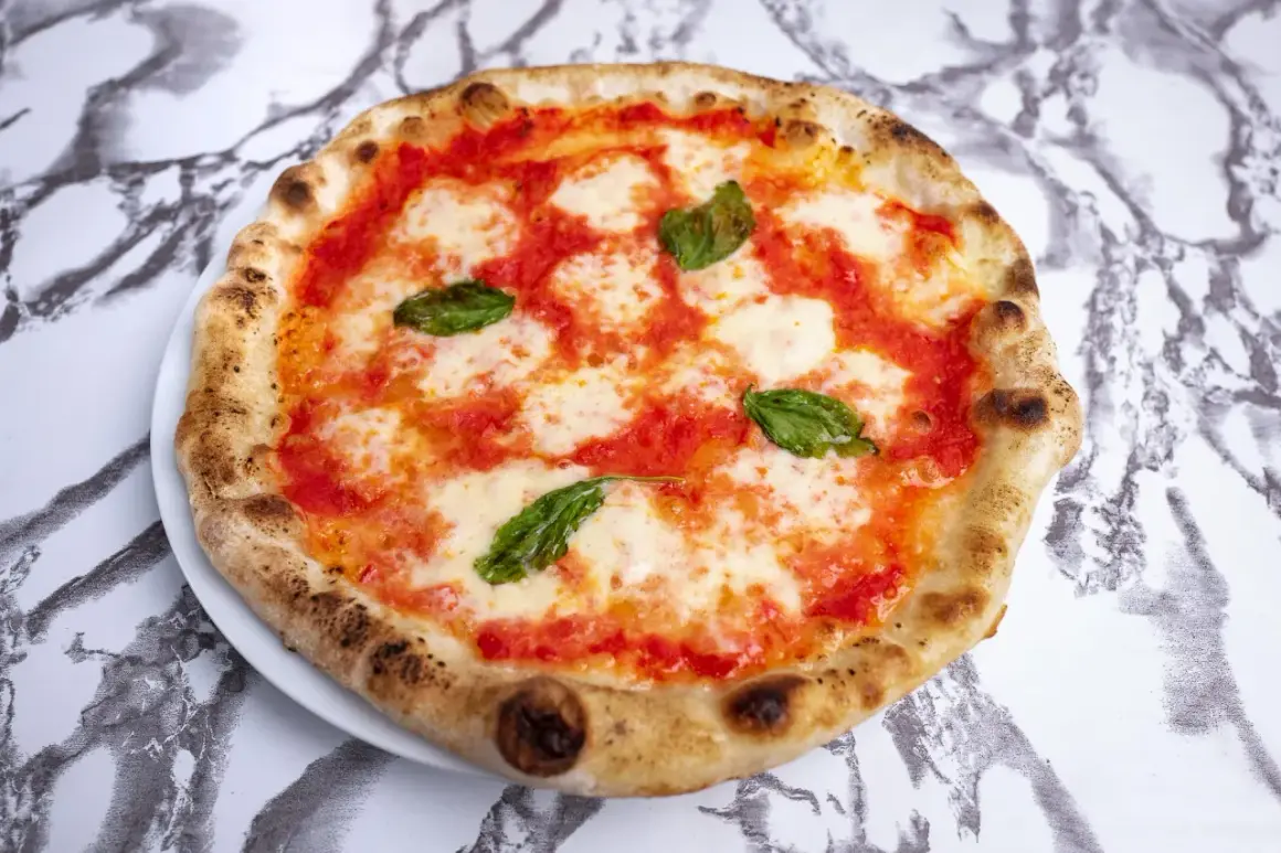 Pyszne ciasto na pizzę neapolitańską, z sosem pomidorowym, mozzarellą i listkami bazylii, na marmurowym tle.