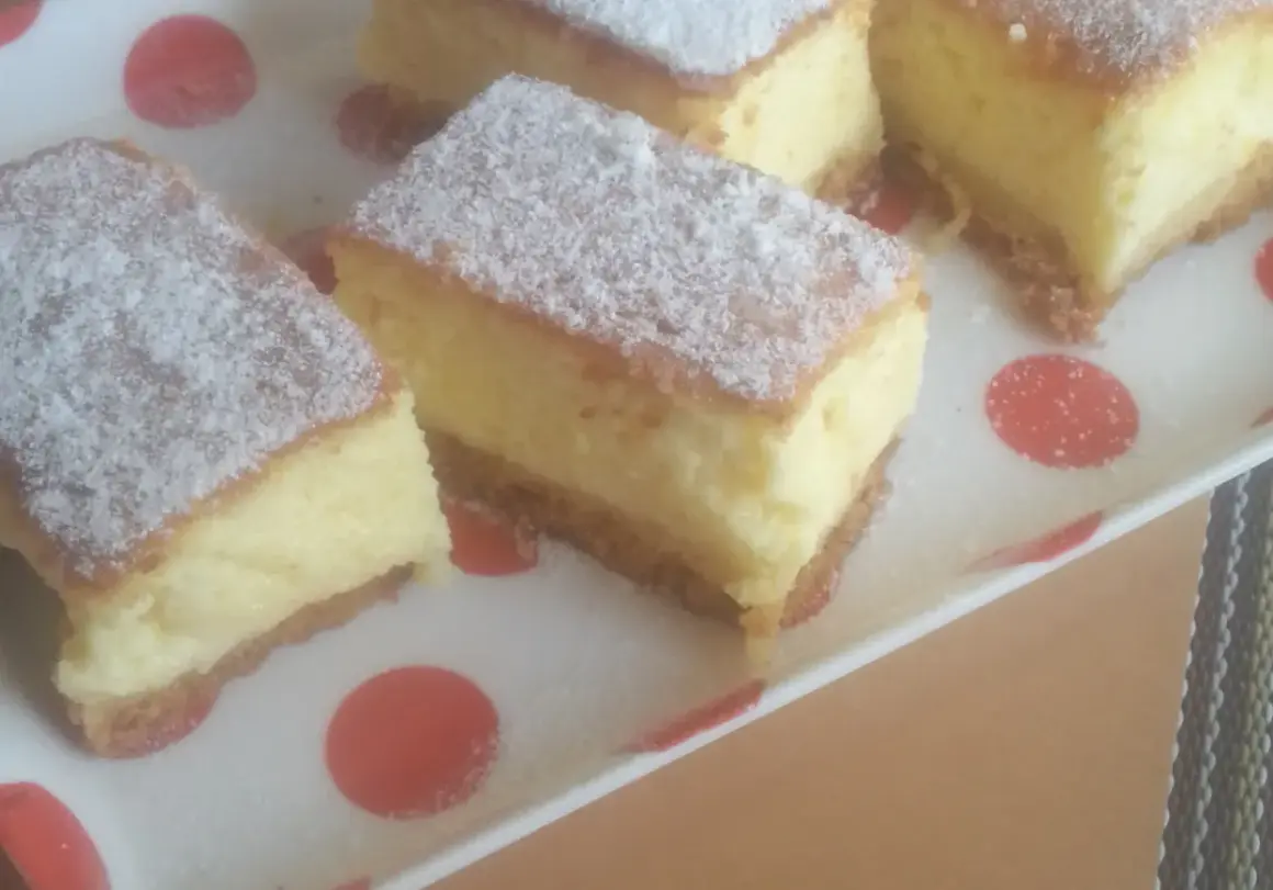 Kawałki puszystego sernika bez sera, posypane cukrem pudrem, na talerzu w czerwone kropki.