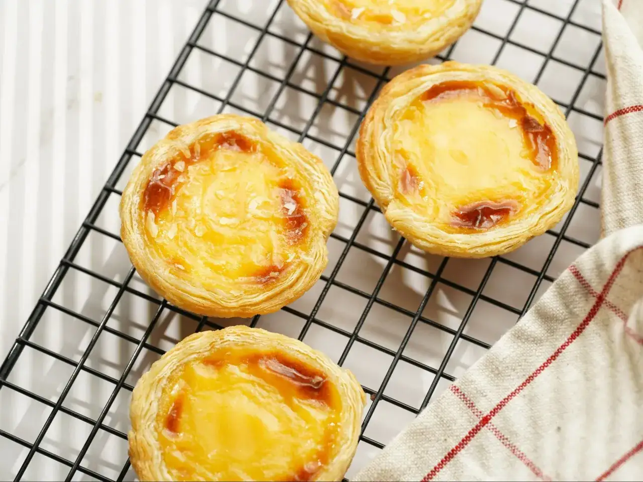 Chrupiące babeczki pastel de nata z kremowym nadzieniem, lekko przypieczone na wierzchu, stygną na czarnej kratce.