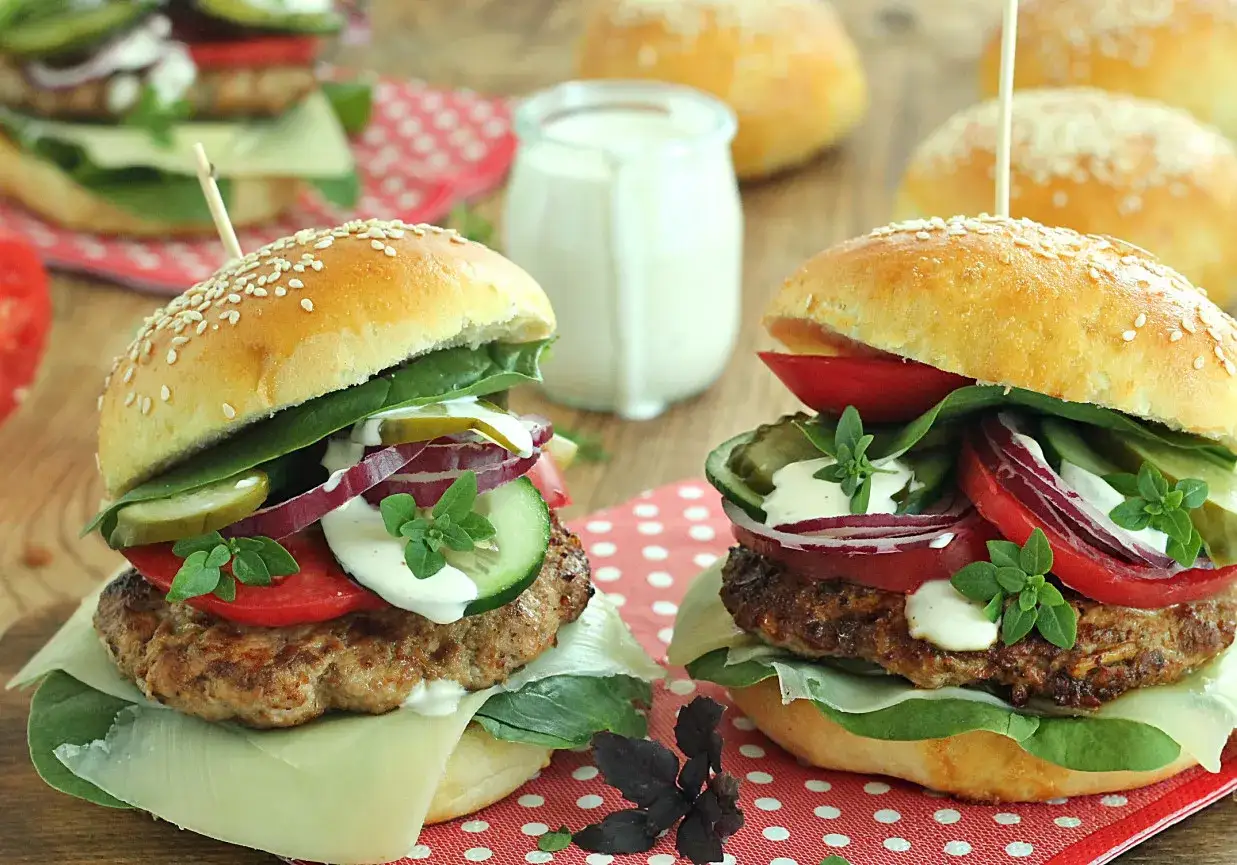 Dwa soczyste burgery z serem, pomidorem, og&oacute;rkiem, czerwoną cebulą i ziołami. Idealne co do burgera, z sosem w słoiczku w tle.