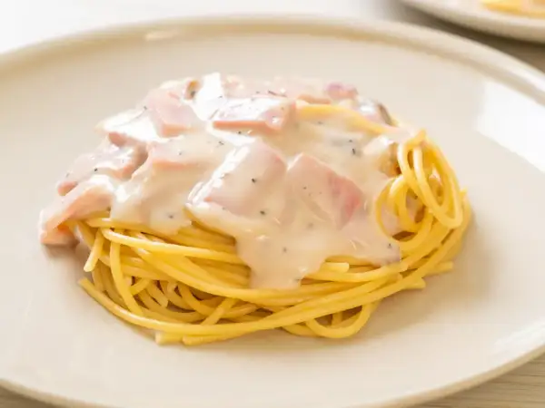 Pyszny makaron z jajkiem i boczkiem w kremowym sosie, posypany pieprzem, podany na białym talerzu.