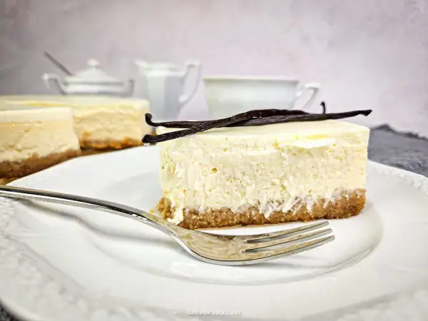 Kawałek puszystego new york cheesecake z wanilią na kruchym spodzie, udekorowany laskami wanilii. Obok widoczne inne serniki i filiżanka.