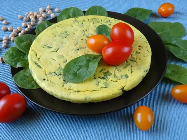 Puszysty omlet z ciecierzycy ze szpinakiem, ozdobiony pomidorkami koktajlowymi i świeżym szpinakiem.