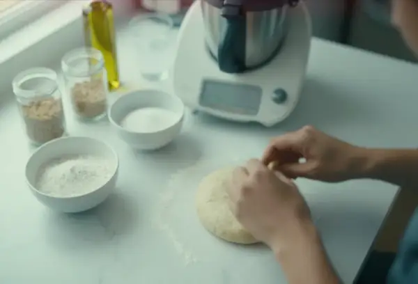 Ręce formują puszyste ciasto na pizzę na blacie. Obok miski z mąką, cukrem i nasionami.