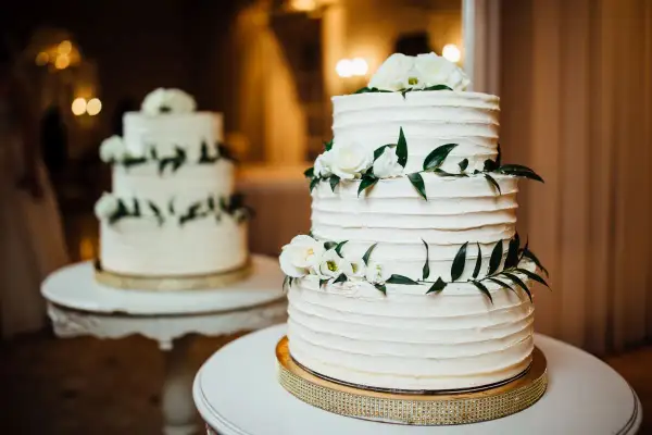 Elegancki tort piętrowy na stelażu, ozdobiony białymi różami i zielonymi liśćmi. Idealny pomysł, jak zrobić tort piętrowy na stelażu na specjalną okazję.